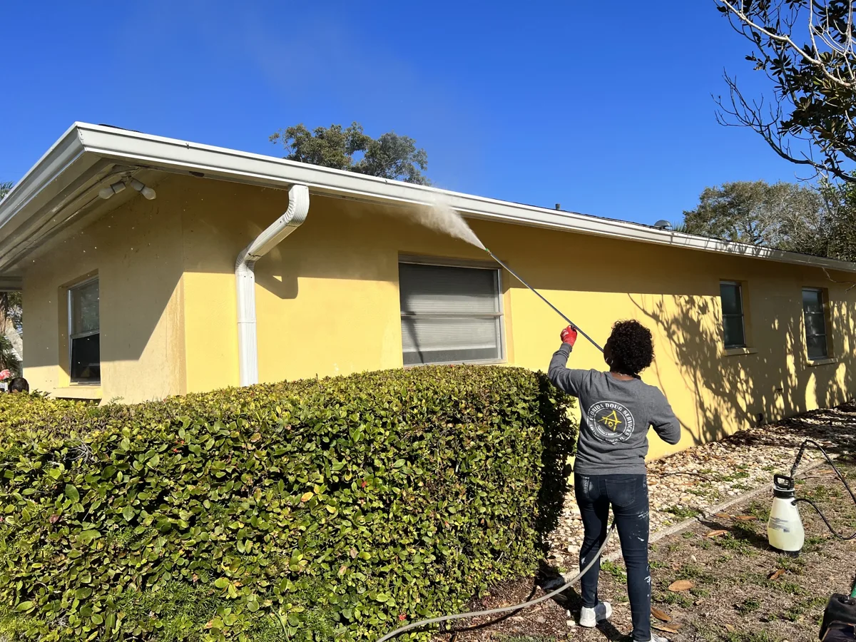 Florida Doug Services crew member soft-washing the stucco walls and gutters of a yellow home