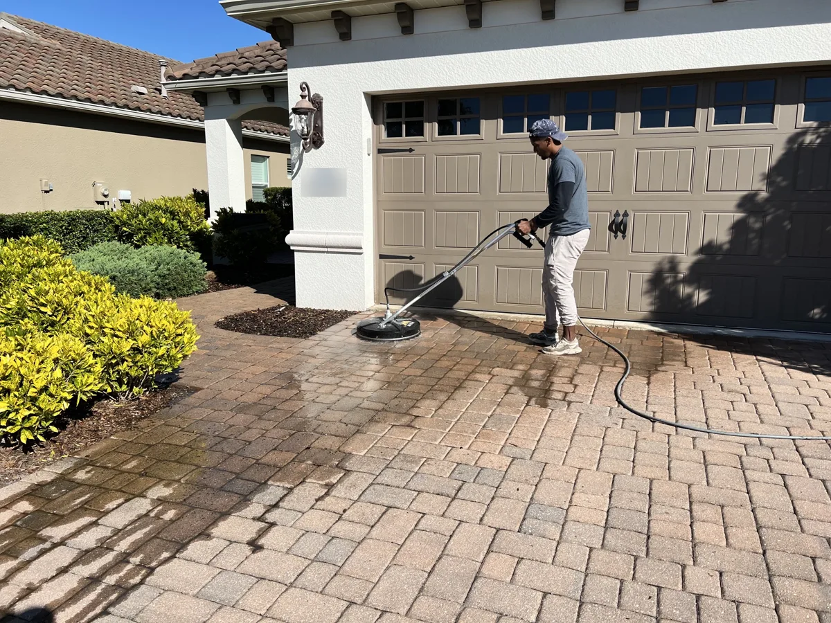 Crew member cleaning a paver driveway with a surface cleaner, showing the clean and dirty sections side by side