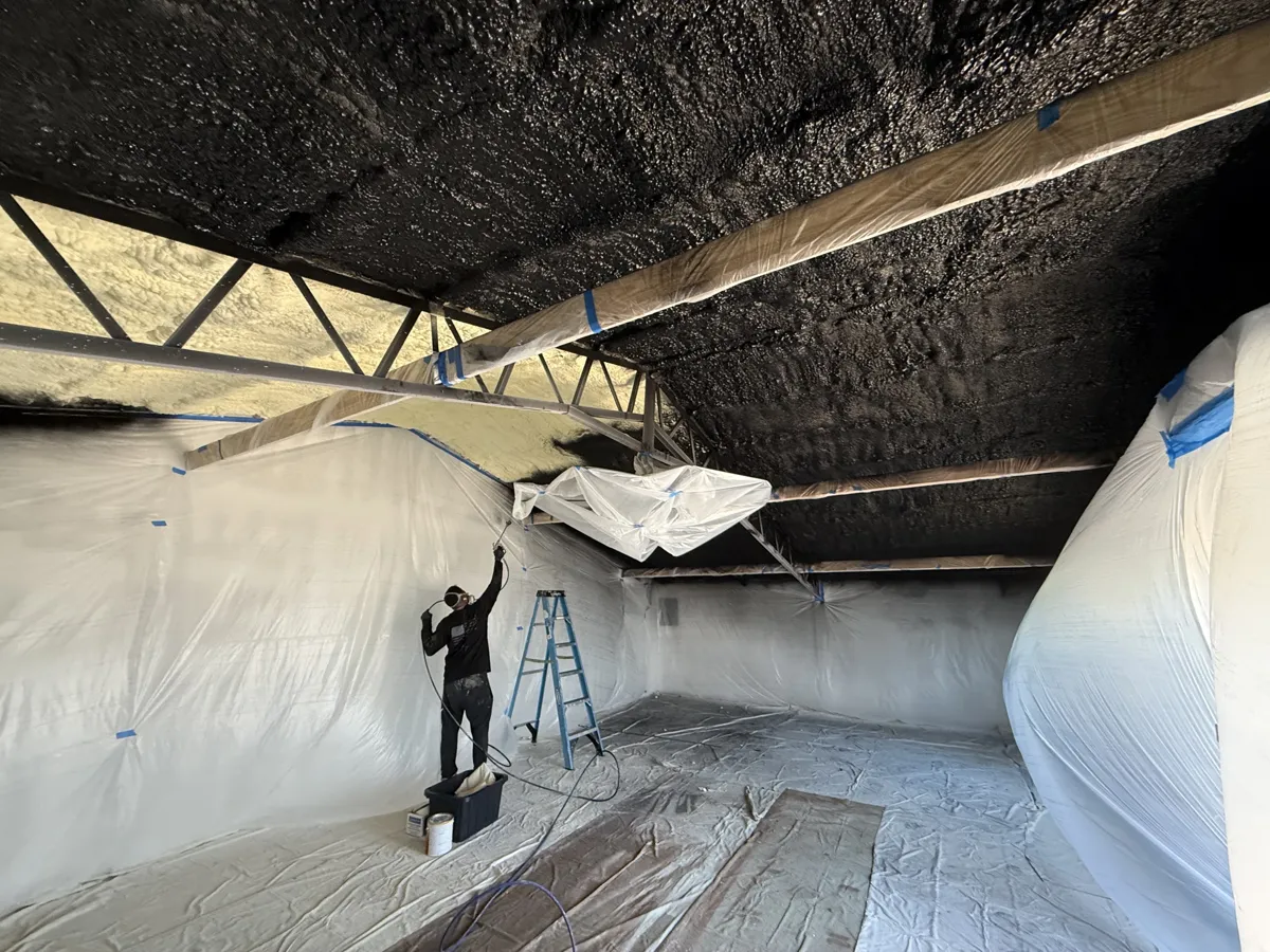 During: room fully wrapped in plastic while a painter sprays the foam ceiling black
