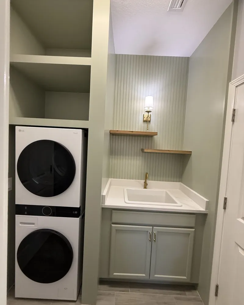 After: sage green laundry room with a built-in surround and open shelves over a stacked washer and dryer, beadboard wall, sink cabinet, floating wood shelves and brass sconce