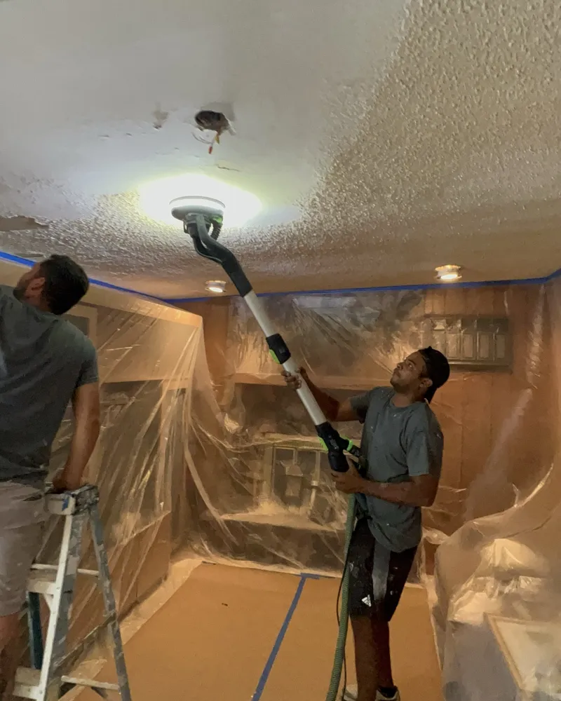 Room fully wrapped in plastic and floor protected while the crew sands popcorn texture off the ceiling with a Festool Planex and dust extractor