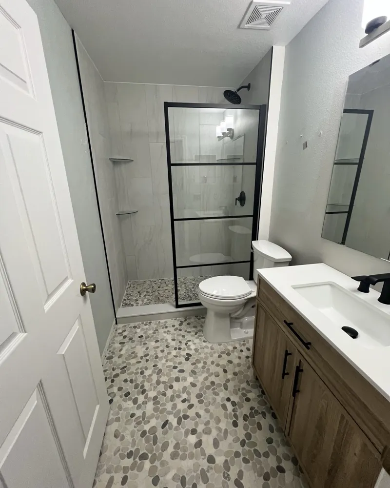 After: walk-in shower with large porcelain wall tile, pebble floor, black-framed glass panel, new wood vanity and matte black fixtures