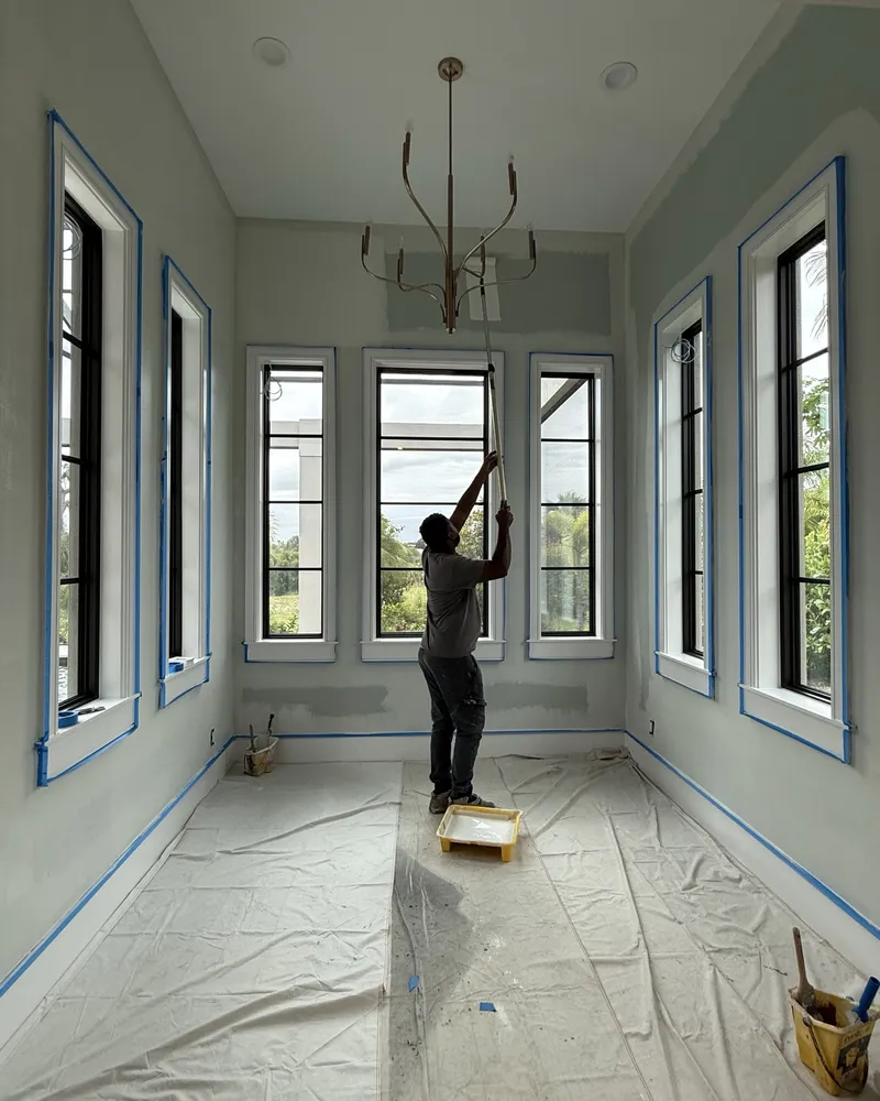 During: painter rolling the ceiling of a bright room with tall black-framed windows, floors covered and trim masked