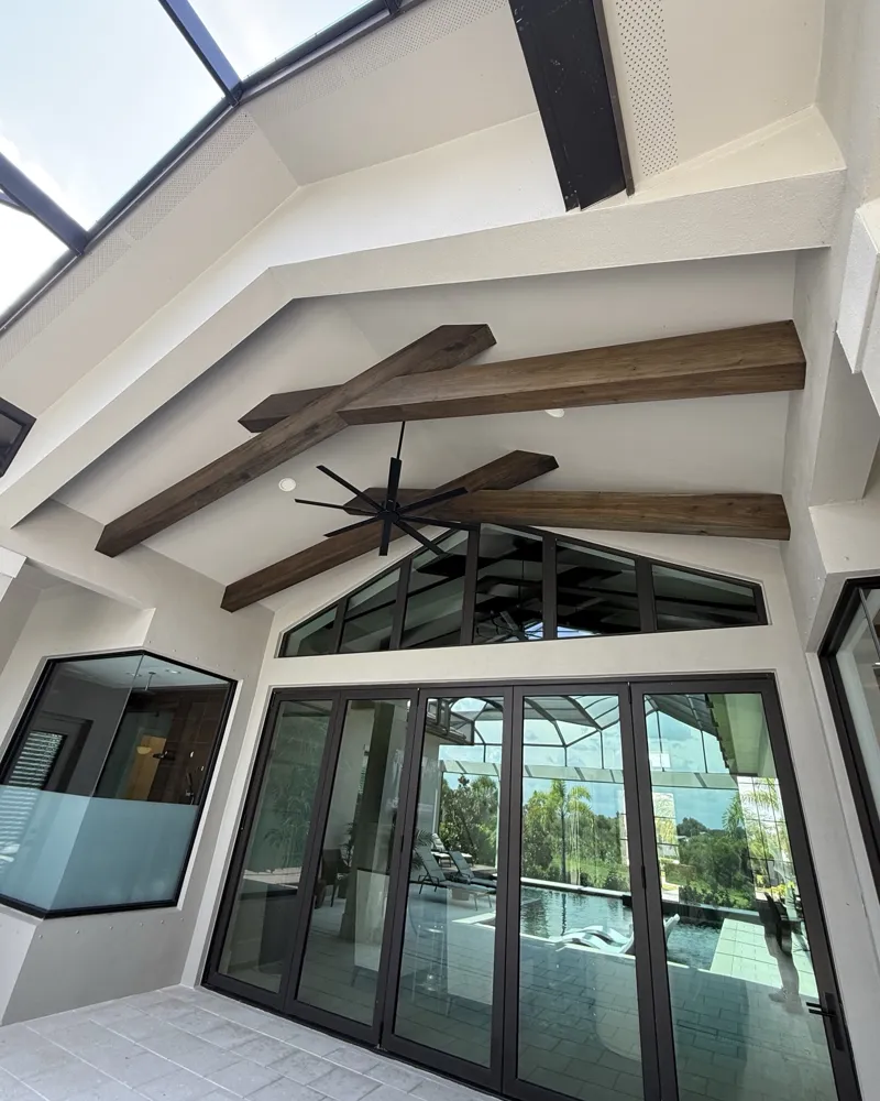 Vaulted lanai ceiling with crossed wood beams sealed in a warm brown finish above folding glass doors