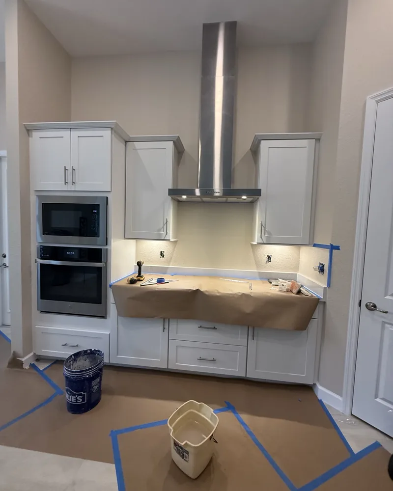 Before: bare textured wall between white shaker cabinets under a stainless range hood, countertop protected for tile work