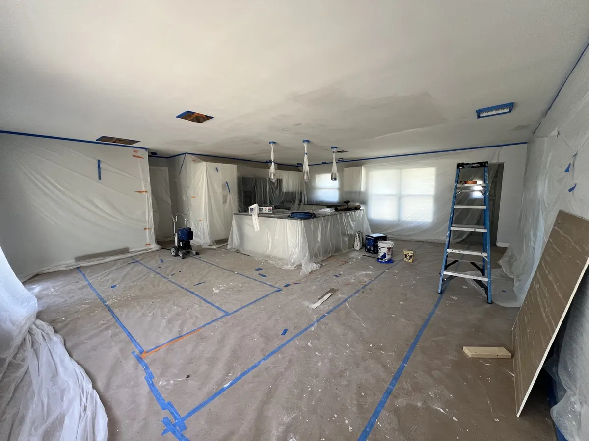 During: open kitchen and living area fully masked in plastic, with ceiling drywall patches and a sprayer ready