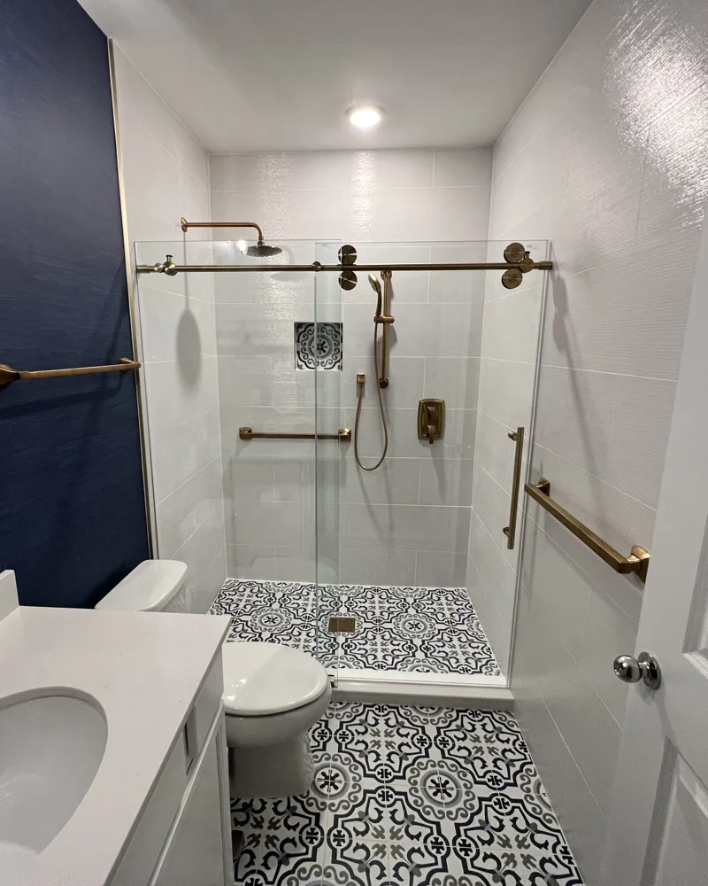 After: walk-in tiled shower with frameless sliding glass door, patterned blue-and-white floor tile, navy grasscloth accent wall and brushed-gold fixtures