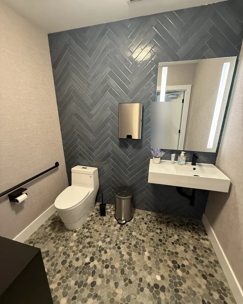After: modern restroom with a navy herringbone tile accent wall, river-pebble floor, textured wallcovering, floating sink and lighted mirror